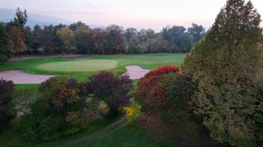 İnsanlar sonbaharda yeşil bir alanda golf oynarlar. Bazı ağaçlarda sarı-kırmızı yapraklar. Delikler için yerler. Bir golf arabası var. Evler ve dağlar sisli bir mesafeden görülebilir. Gün batımı. Düğün.