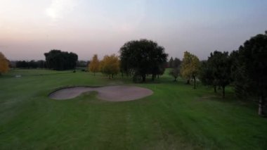 Bir kuş sürüsü golf sahalarının üzerinde uçuyor. Yeşil tarlalardan, sarı-yeşil sonbahar ağaçlarından ve günbatımından gelen hava manzarası. Delikler için ayrılan yerler var. Parkta birkaç gölet var. Almaty, Kazakistan