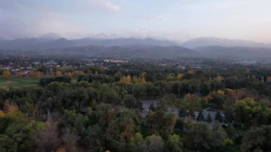 Karlı tepeler manzaralı sonbahar golf parkı. Altın ağaçların, sarı-kırmızı yaprakların ve yeşil otların hava manzarası. Evler ve dağlar uzaktan görülebilir. Gün batımı. Kazakistan