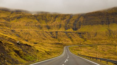 Streymoy adasındaki tepeler boyunca asfalt yol. Vadide kalın bulutlar var. Faroe Adaları, Danimarka.