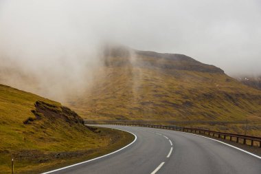 Streymoy adasındaki tepeler boyunca asfalt yol. Vadide kalın bulutlar var. Faroe Adaları, Danimarka.