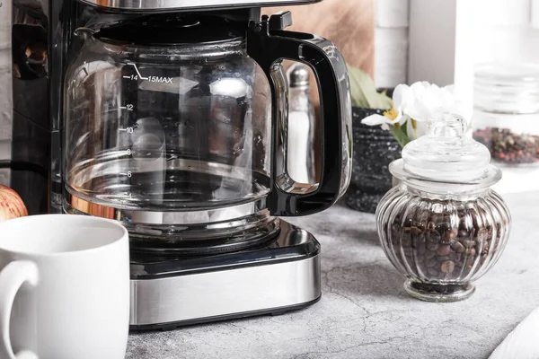 Una macchina da caffè elettrica a goccia nera con una teiera di vetro prepara una bevanda mattutina. Elettrodomestici, una tazza bianca e un barattolo di fagioli sul tavolo della cucina sul piano di lavoro a casa Foto Stock Royalty Free