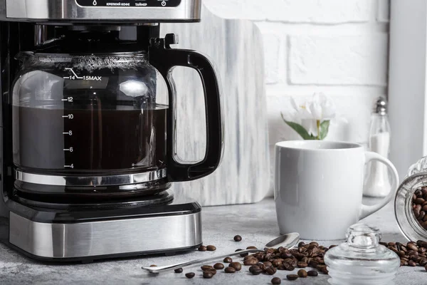 Чорна крапельна електрична кавоварка зі скляним чайником заварює ранковий напій. Побутова техніка, біла чашка і баночка з бобами на кухонному столі на стільниці вдома Стокове Зображення
