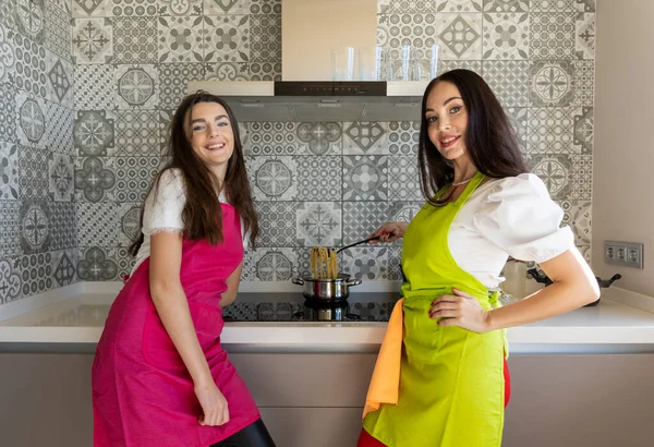 在家里的现代厨房里 穿着围裙的快乐的年轻女性和成年女性一边微笑着 一边看着相机 一边煮着意大利面 — 图库照片