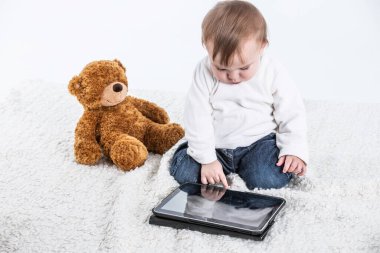 Bir tabletin ekranına dokunan bir bebeğin beyaz arkaplanlı fotoğrafı ve yanında oyuncak ayı ve içecek saplı bir kap..