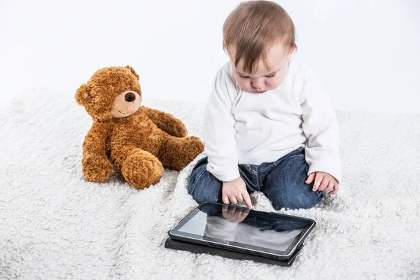 Bir tabletin ekranına dokunan bir bebeğin beyaz arkaplanlı fotoğrafı ve yanında oyuncak ayı ve içecek saplı bir kap..