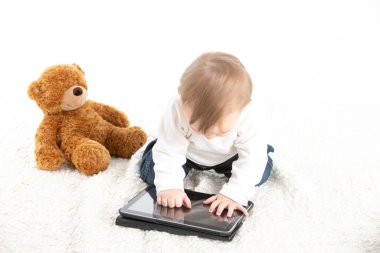 Yanında oyuncak ayı olan bir tabletin ekranına dokunan bir bebeğin beyaz arkaplanlı fotoğrafı.