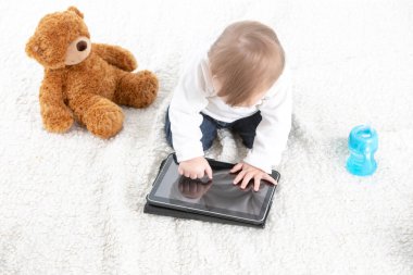 Beyaz arkaplanlı, yanında oyuncak ayıyla tabletin ekranına dokunan bir bebek resmi ve içecek saplı bir kap..