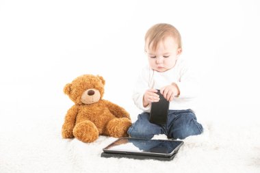 Elinde cep telefonu olan, tabletli ve yerde oyuncak ayısı olan bir bebeğin beyaz arka plan fotoğrafı..