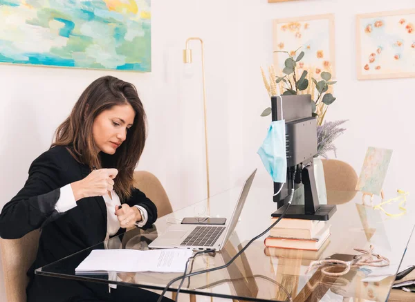 Female manager in formal outfit working with laptop and making notes on paper at table with computer monitor and medical mask and doing telework from home during coronavirus pandemic
