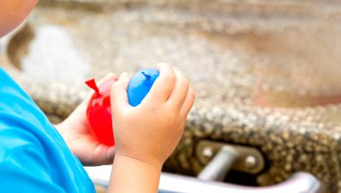 Çocuk parkın çeşmesinde iki su balonuyla oynuyor. Çocuğun kırmızı ve mavi bir su balonu var..