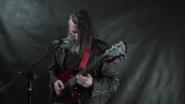 Músico de rock barbudo em jaqueta de couro toca guitarra elétrica vermelha e canta no palco com fundo preto — Vídeo de Stock