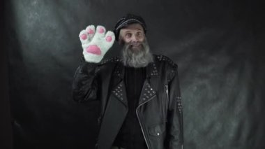 Deri ceketli ve şapkalı sakallı rock yıldızı siyah perde arkaplanlı gece kulübü konserinde beyaz kedi pençesi eldiveniyle seyirciye gülümsüyor ve el sallıyor.