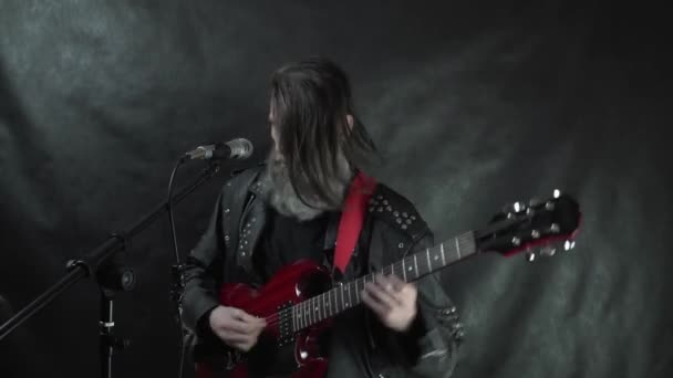 लेदर जॅकेटमध्ये लांब केसांचा रॉक संगीतकार लाल इलेक्ट्रिक गिटार वाजवतो आणि काळ्या पार्श्वभूमीसह स्टेजवर गातो — स्टॉक व्हिडिओ
