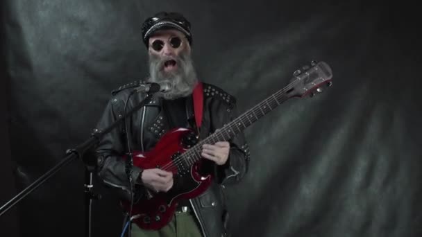 Musicien rock barbu en veste et casquette en cuir portant des lunettes rondes joue de la guitare électrique rouge et chante sur scène avec un fond noir — Video