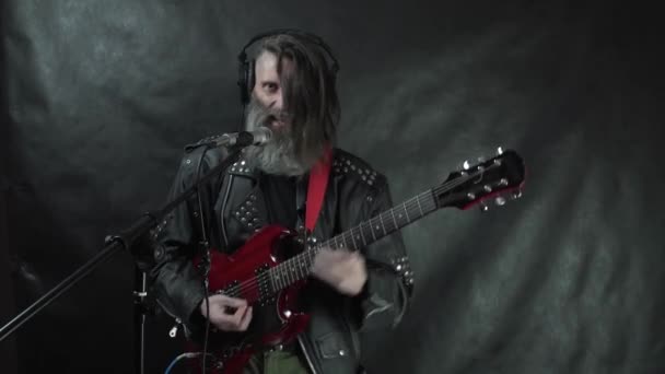 Бородатий рок-музикант в навушниках з голими храмами і шкіряною курткою грає на червоній електрогітарі, співає і записує свою композицію в студії — стокове відео