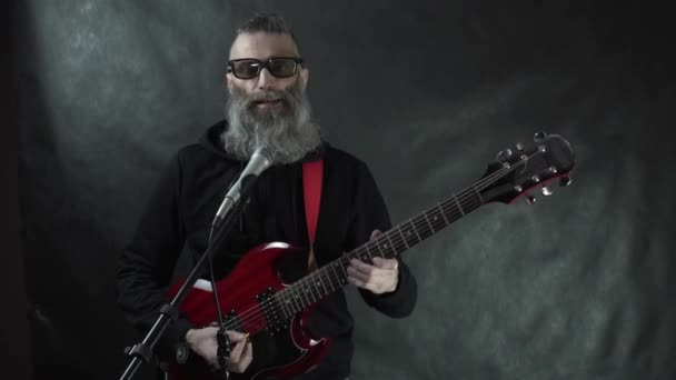 Bärtiger haariger Rockstar mit Retro-Sonnenbrille und schwarzem Kapuzenpulli mit rasierten Schläfen spielt auf roter E-Gitarre auf der Bühne mit schwarzem Hintergrund — Stockvideo