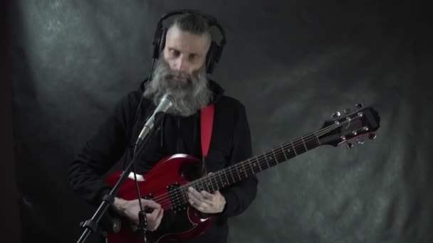 Musicista rock barbuto con cuffie, templi rasati e cappuccio nero suona la chitarra elettrica rossa, canta e registra la sua composizione in studio — Video Stock