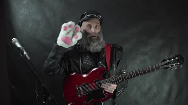 Rockstar barbudo con chaqueta de cuero, guitarra eléctrica roja y gorra sonriendo y saludando a la audiencia con guante de pata de gato blanco en concierto de discoteca con fondo de cortina negro — Vídeos de Stock