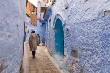 Fas 'taki Chefchaouen Medina' da. Geleneksel Berberi paltosu giymiş bir adam mavi duvarlarla kaplı bir sokakta yürüyor.