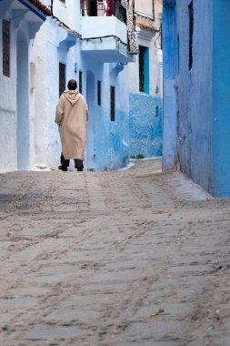 Fas 'taki Chefchaouen Medina' da. Geleneksel Berberi paltosu giyen bir adam mavi cepheli bir sokakta yürüyor..