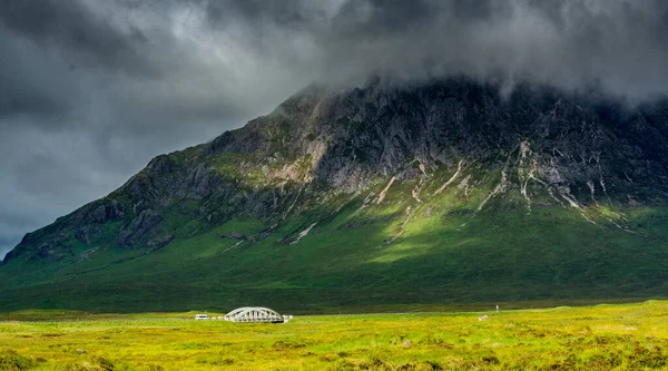Batı Highland Yolu boyunca. Stob a Ghlais Choire 'ın eteğindeki Glen Coe vadisinde bir nehir boyunca uzanır. Gökyüzü tehditkar ve güneş dağın duvarlarında oynuyor.