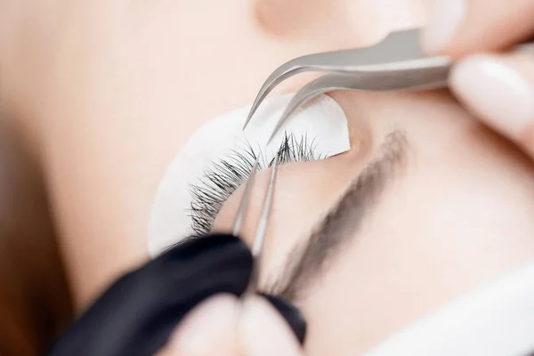 Closeup Eyelash extension procedure. Master tweezers fake black long lashes beautiful female eyes — Stock Photo, Image