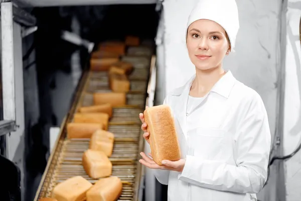 パンを手に新鮮なパンを持っているベイカーの女性は、食品パン工場の背景自動コンベア — ストック写真