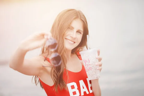 Happy sourire femme en maillot de bain rouge tient verre avec cocktail sur la plage sur des lunettes, la lumière du soleil — Photo