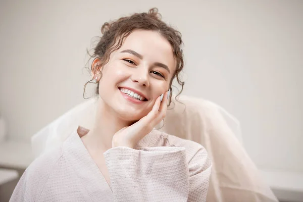 Νεαρή όμορφη γυναίκα με σγουρά μαλλιά χαμόγελα και κοιτάζει στο πλαίσιο σε ρόμπα, έννοια της αισθητικής σαλόνι ομορφιάς — Φωτογραφία Αρχείου