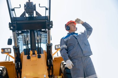 Buldozer önünde miğferli bir adam ya da inşaat alanında kazıcı. Teknoloji ve sanayi işçilerinin etkileşimi kavramı
