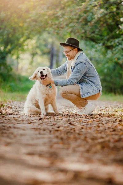 Понятие дружбы между животным и человеком. Путешествующий блоггер в шляпе с золотистым ретривером, обнимающий и веселый — стоковое фото