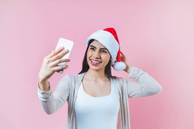 Genç Latin bir kadın cep telefonu tutuyor. Pembe arka planda bir Noel konseptinde fotoğraf çekiyor.