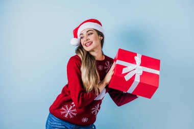 Latin Amerika 'da genç bir kadın Noel hediyelerini mavi arka planda, Noel şapkasıyla tutuyor.