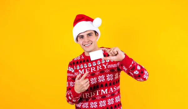 图上的拉丁男子手持空白卡片在圣诞节购物时头戴黄色背景的桑塔帽的肖像 — 图库照片