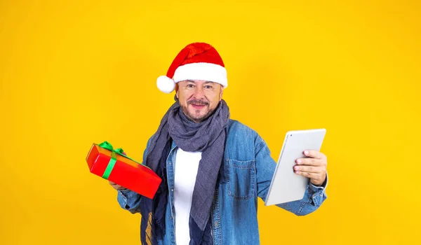 在墨西哥 一位拿着圣诞礼物和带着圣诞帽的数码平板电脑的老年男子 背景是黄色的 — 图库照片