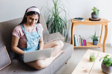 Genç Latin kız öğrenci video konferansında ya da internet dersinde bilgisayar kullanıyor ve Meksika 'da evde kanepede otururken yeşil meyve suyu içiyor. 