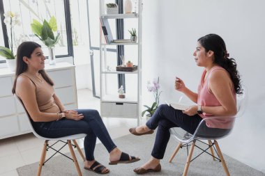 Hispanic young woman psychologist and female patient telling about mental problems while doctor is listening and making notes. Psychotherapy concept in Mexico Latin America