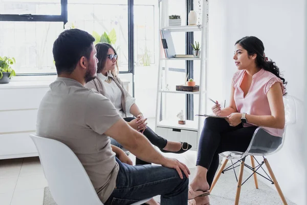 Hispanic young woman psychologist with couple patient in therapy talking about mental problems while doctor is listening and making notes in Mexico Latin America