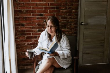 Pensive relaxed latin mature woman reading a book at home and drinking coffee sitting at bedroom in Mexico Latin America
