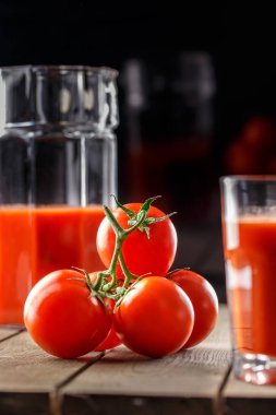 Yanında domates suyu olan cam bir bardak, bir domates dalı uzanıyor. Ahşap bir masanın üzerinde bir bardak domates suyu ve domates var. Siyah bir arka planda yatıyor..