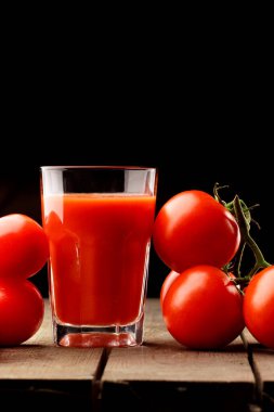 Yanında domates suyu olan cam bir bardak, bir domates dalı uzanıyor. Ahşap bir masanın üzerinde bir bardak domates suyu ve domates var. Siyah bir arka planda yatıyor..