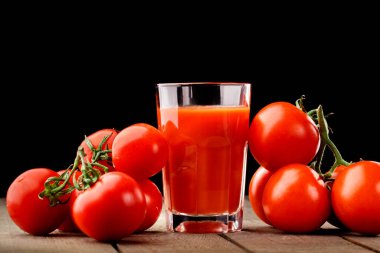 Yanında domates suyu olan cam bir bardak, bir domates dalı uzanıyor. Ahşap bir masanın üzerinde bir bardak domates suyu ve domates var. Siyah bir arka planda yatıyor..