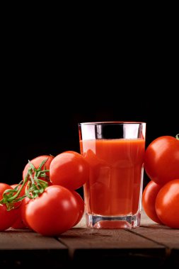 Yanında domates suyu olan cam bir bardak, bir domates dalı uzanıyor. Ahşap bir masanın üzerinde bir bardak domates suyu ve domates var. Siyah bir arka planda yatıyor..
