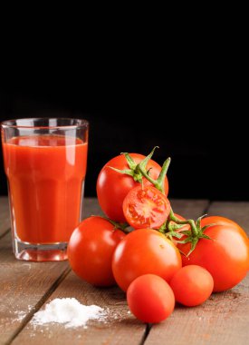 Yanında domates suyu olan cam bir bardak, bir domates dalı uzanıyor. Ahşap bir masanın üzerinde bir bardak domates suyu ve domates var. Siyah bir arka planda yatıyor..