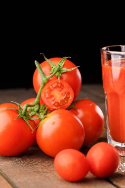 Yanında domates suyu olan cam bir bardak, bir domates dalı uzanıyor. Ahşap bir masanın üzerinde bir bardak domates suyu ve domates var. Siyah bir arka planda yatıyor..