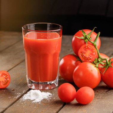 Yanında domates suyu olan cam bir bardak, bir domates dalı uzanıyor. Ahşap bir masanın üzerinde bir bardak domates suyu ve domates var. Siyah bir arka planda yatıyor..
