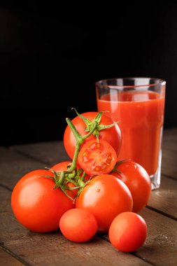 Yanında domates suyu olan cam bir bardak, bir domates dalı uzanıyor. Ahşap bir masanın üzerinde bir bardak domates suyu ve domates var. Siyah bir arka planda yatıyor..