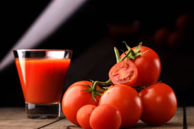 Yanında domates suyu olan cam bir bardak, bir domates dalı uzanıyor. Ahşap bir masanın üzerinde bir bardak domates suyu ve domates var. Siyah bir arka planda yatıyor..