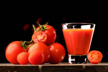 Yanında domates suyu olan cam bir bardak, bir domates dalı uzanıyor. Ahşap bir masanın üzerinde bir bardak domates suyu ve domates var. Siyah bir arka planda yatıyor..